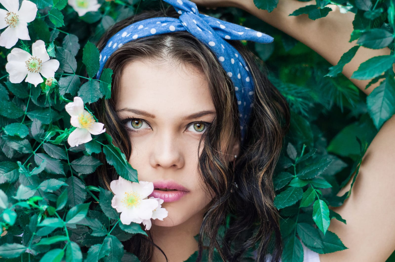 a closeup portrait of a woman with dark wavy hair wearing a blue polka dot headband, surrounded by lush green leaves and white flowers